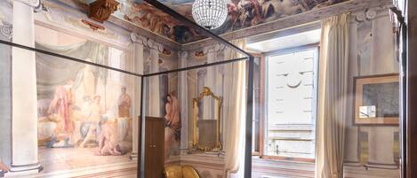 A room with a bed, a golden chair, a large mirror, and a ceiling mural.
