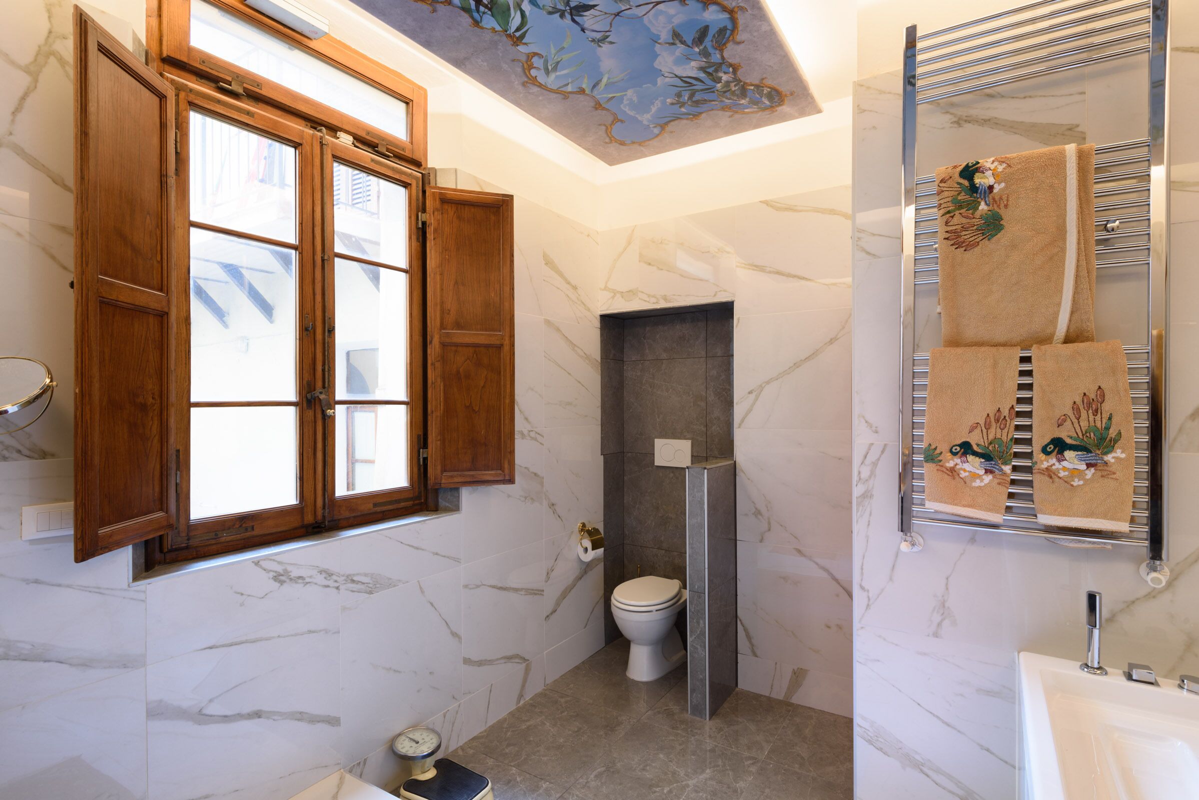 A bathroom with a marble wall, a wooden window, a towel rack with towels, and a toilet.