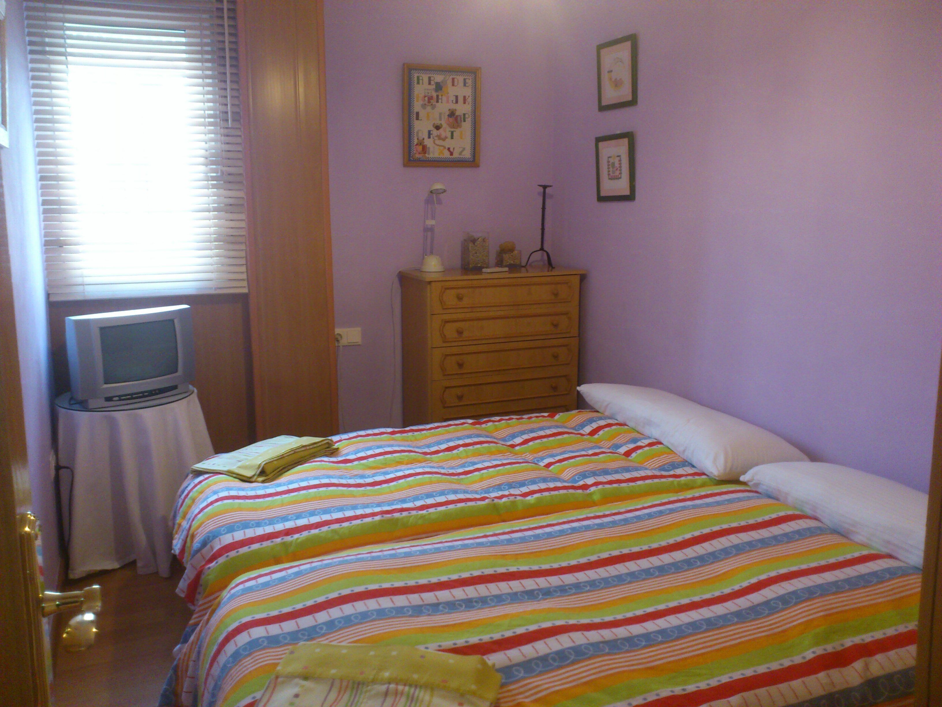 Un dormitorio con cama, cómoda, televisor sobre una mesita y ventana con persianas.