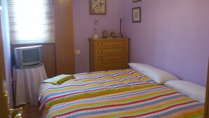 Une chambre à coucher avec un lit, une commode, un téléviseur posé sur une petite table et une fenêtre avec des stores.