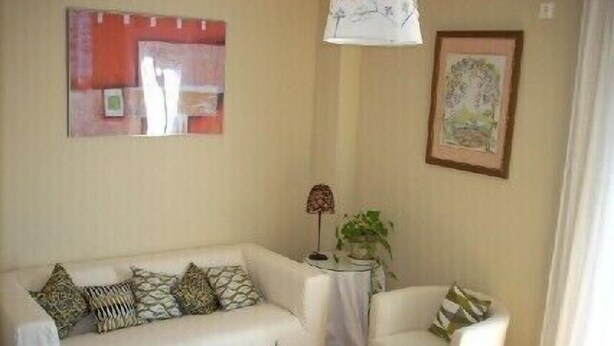 Un salon avec un canapé blanc, un fauteuil blanc, une petite table et une lampe. Un tableau encadré est accroché au mur et une plante est posée sur une table d’appoint.