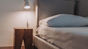 Ein ordentlich bezogenes Bett mit Kopfstütze, einer Nachttischlampe und einem kleinen Holfnachtkästchen.