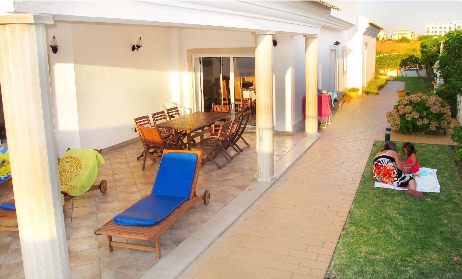 Une terrasse couverte avec une table et des chaises, une chaise longue et un petit jardin où deux personnes sont assises sur l’herbe.