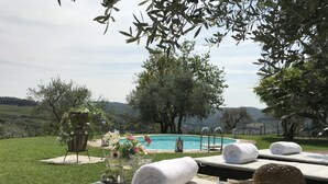 A poolside area with a view of a swimming pool, a grassy lawn, and trees. There are lounge chairs with towels and a hat, and a table with a vase of flowers.