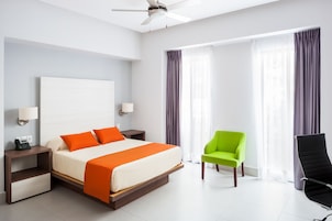 Una habitación de hotel con una cama, una silla verde, una silla negra, una mesita de noche y un ventilador de techo.