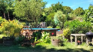 Un jardín con piscina azul, un carretón de madera y una parrilla de barbacoa.
