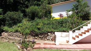 Un jardin avec un mur en pierre, un escalier et un bâtiment à la façade jaune et blanche.