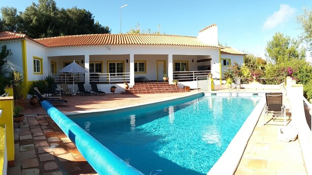 A rectangular swimming pool with a blue edge, surrounded by a patio area with lounge chairs and a covered seating area. - Quinta da princesa Barbara (Caldas da Rainha)