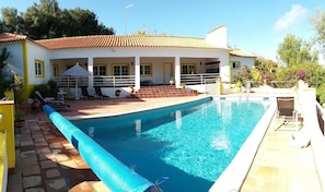 Une piscine rectangulaire avec un bord bleu, entourée d’une terrasse aménagée avec des chaises longues et un espace salon couvert.