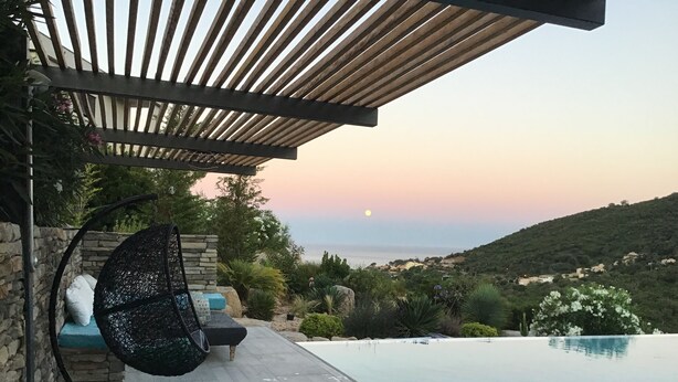 Area a bordo piscina con una sedia sospesa, un muro in pietra e vista sul mare al tramonto.