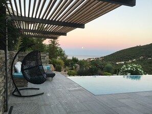 Zwembadterras met een hangstoel, een stenen muur en uitzicht op zee bij zonsondergang.