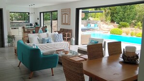 Un soggiorno spazioso con divano, poltrone e tavolo da pranzo. È presente un angolo cottura con tavolo e sedie. Un'ampia finestra offre vista su piscina e giardino esterno.