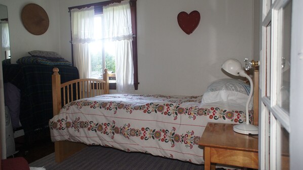 Une chambre à coucher avec un lit, une table de chevet en bois, un bureau, une chaise, une fenêtre avec des rideaux et un objet décoratif en forme de cœur accroché au mur.