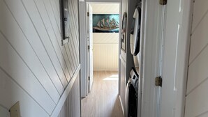 Un pasillo estrecho con paneles blancos, una puerta a la derecha y vista a una habitación con piso de madera y un cuadro en la pared.