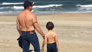 Un hombre y un niño caminando sobre una playa arenosa con olas del océano al fondo.