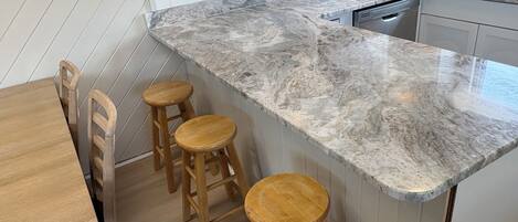 A kitchen with a marble countertop, wooden stools, and a dishwasher.