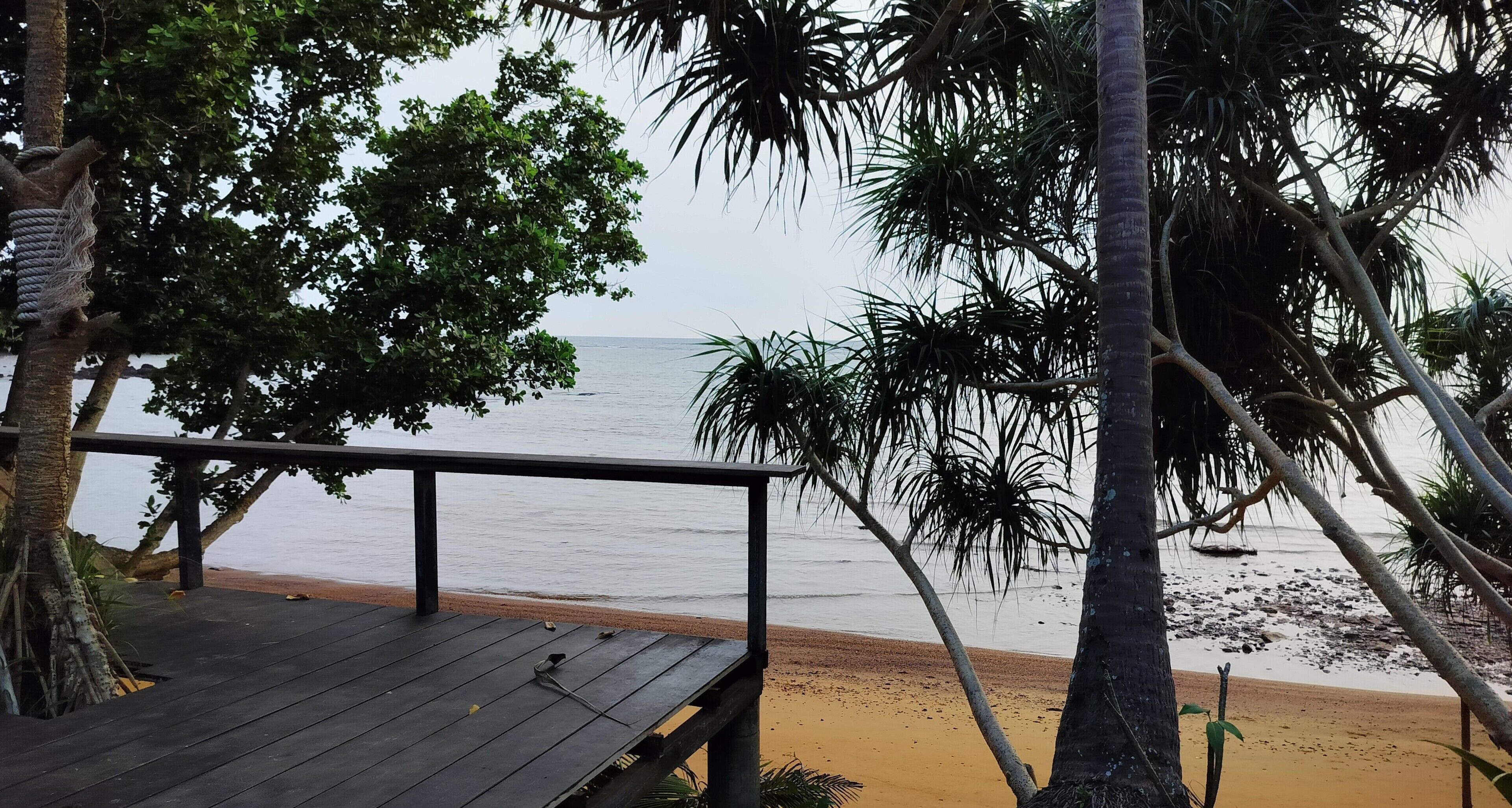 Eine Holzterrasse mit Blick auf einen Strand, Palmen und das Meer.