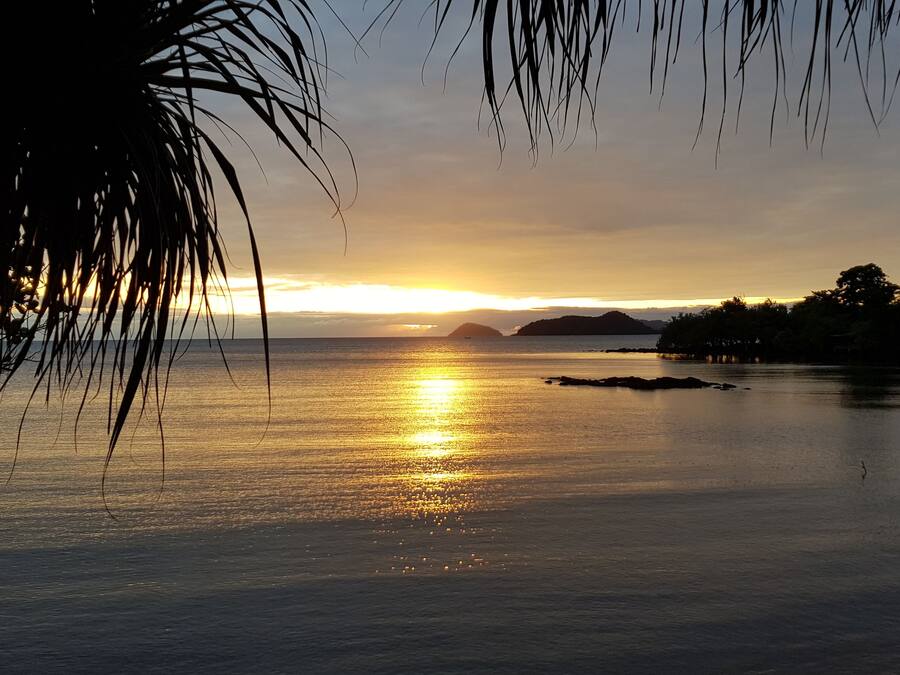 Ein Sonnenuntergang über einem ruhigen Meer, im Hintergrund Landformen und Palmen, die den Blick einrahmen.