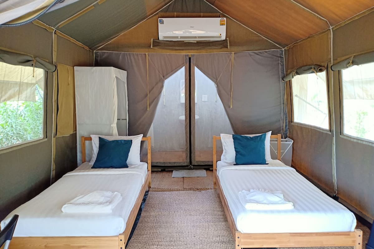 Un hébergement sous tente comprenant deux lits simples, chacun avec un oreiller et une serviette pliée. Un appareil de climatisation est fixé au plafond.