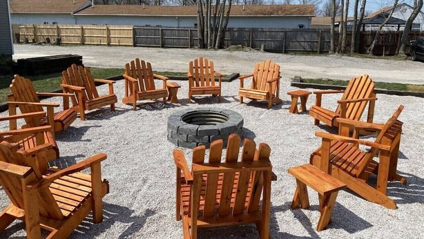 Un ensemble de chaises Adirondack en bois disposées en cercle autour d’un foyer central, dans un espace recouvert de gravier, avec une clôture en bois et des bâtiments en arrière-plan.