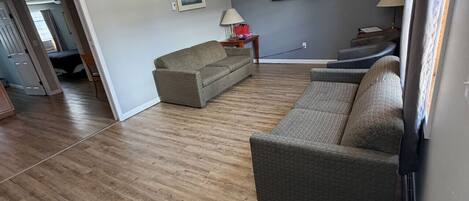 A spacious living room with a wooden floor, two sofas, a TV, and ceiling fans.