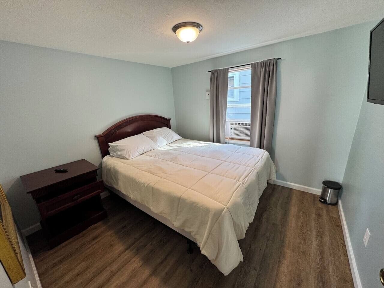 A bedroom with a bed, a nightstand, a small trash bin, and a window with curtains.