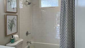 A bathroom with white tiled walls, a bathtub, a showerhead, and a window. - A Great Apartment In The Middle Of It All! (Nashville)