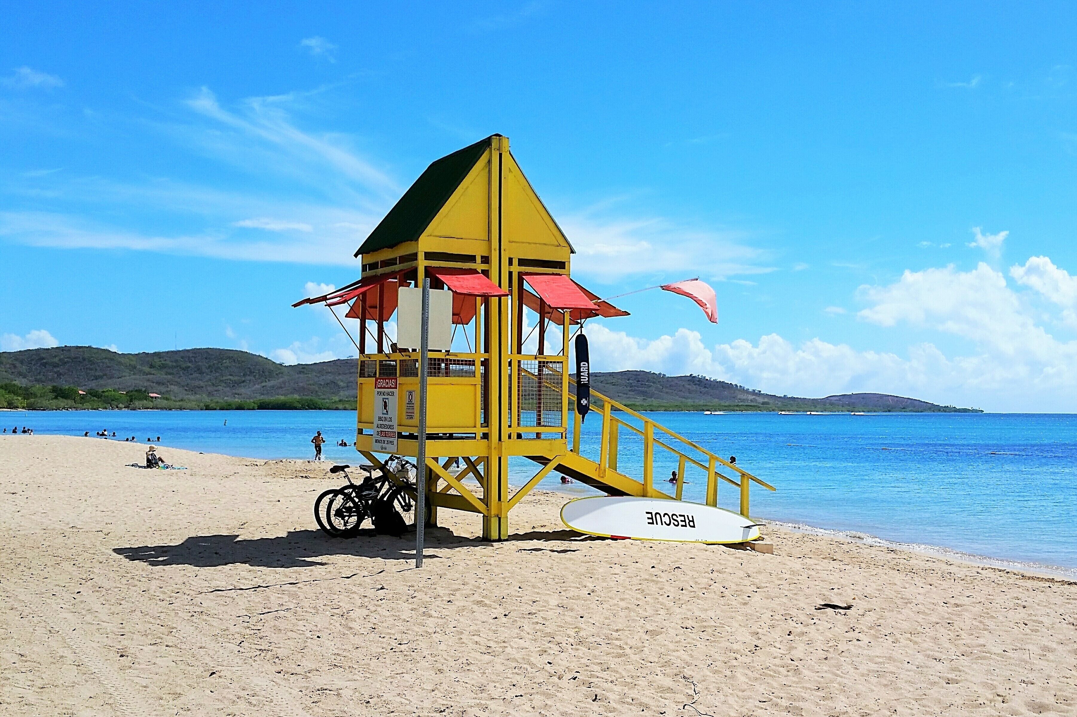 Una torre de salvavidas con un esquema de color rojo y amarillo, ubicada en una playa arenosa con agua azul cristalina y colinas al fondo.