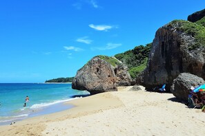 Une plage avec une eau d’un bleu limpide, une grande formation rocheuse et quelques personnes.