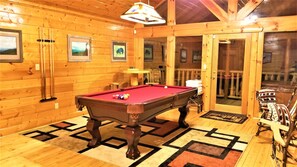 A wooden interior with a pool table, framed pictures, and a view to the outside.