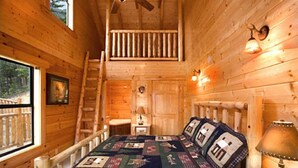 A wooden cabin bedroom with a bed, a nightstand, a lamp, a ladder, and a window with a view of the outdoors. - Aug Special-Mountain View, 2 Suites, Hot Tub, Game Rm, Hammock, Fire Pit + More! (Sevierville)