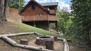 A wooden cabin with a balcony, surrounded by a natural landscape with a fire pit area. - Aug Special-Mountain View, 2 Suites, Hot Tub, Game Rm, Hammock, Fire Pit + More! (Sevierville)