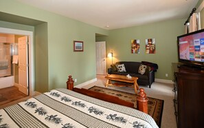 Un dormitorio con cama, mesita de noche de madera, escritorio, silla, sofá, televisor y puerta que da a un baño.