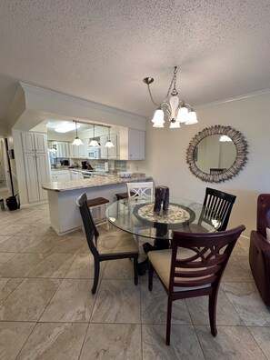 A dining area with a glass table, dark wooden chairs, a round mirror, and a kitchen in the background.