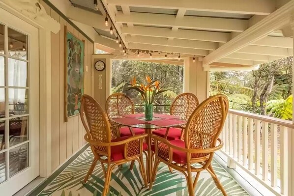 A covered outdoor dining area with wicker chairs and a glass table, surrounded by lush greenery.
