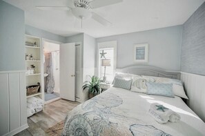 Un dormitorio con cama, ventilador de techo, mesita de noche, lámpara y estante con toallas.
