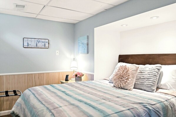 A hotel room with a bed, a nightstand, a lamp, and a welcome sign on the wall.