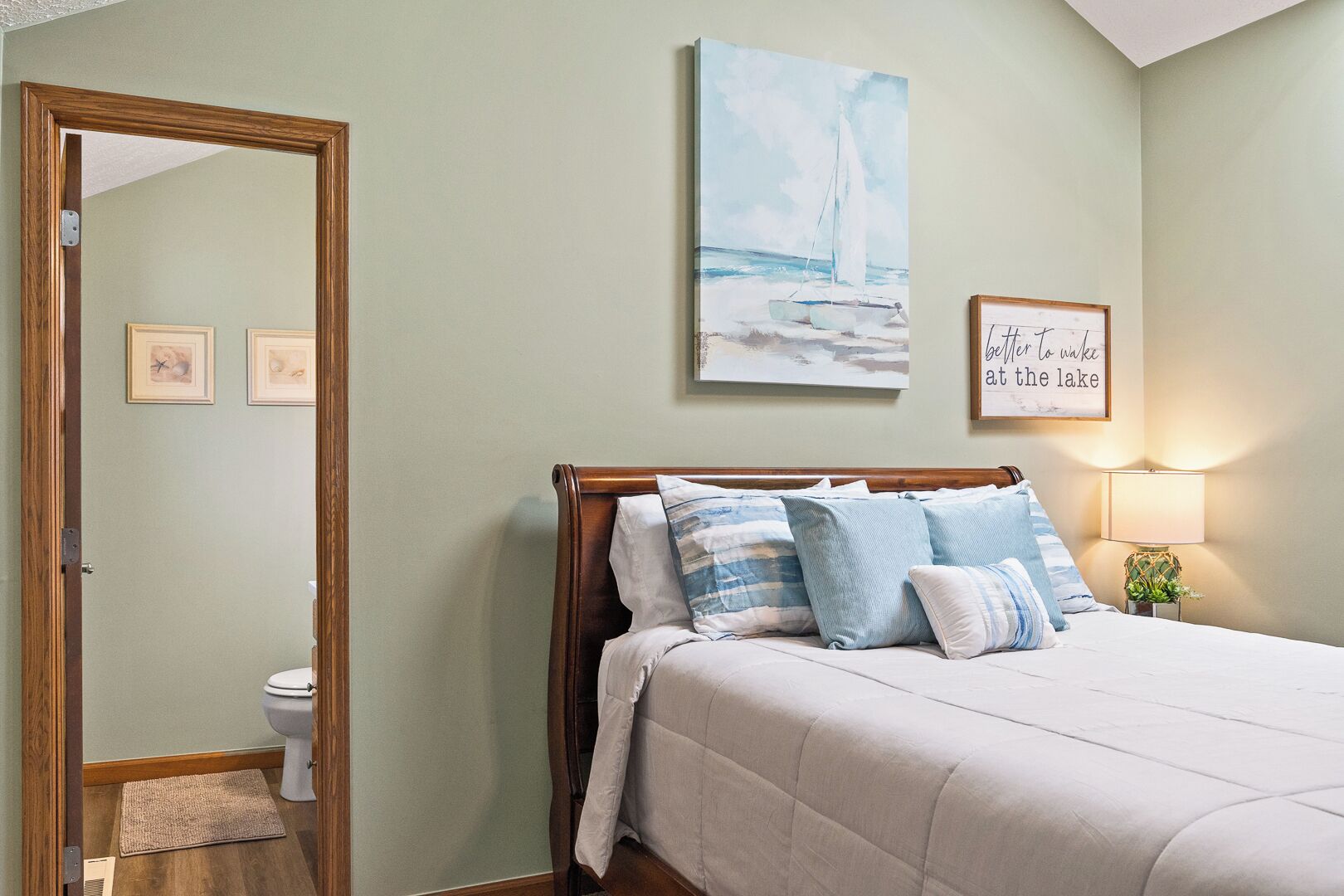 A bedroom with a bed, a bedside table with a lamp, a painting, and a framed quote on the wall.