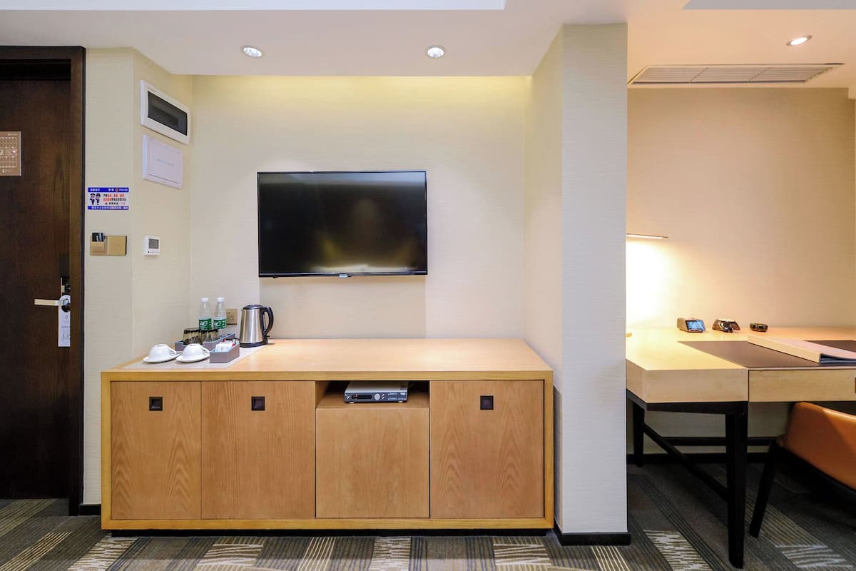 A hotel room with a wooden cabinet, a flat-screen TV mounted on the wall, a coffee maker, and a desk with a lamp.
