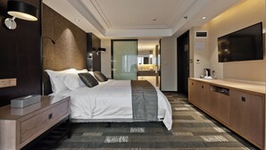 Ein modernes Hotelzimmer mit einem großen Bett, Nachttischen, einem Flachbildfernseher und einem durch eine Glastür einsehbaren Badezimmer.