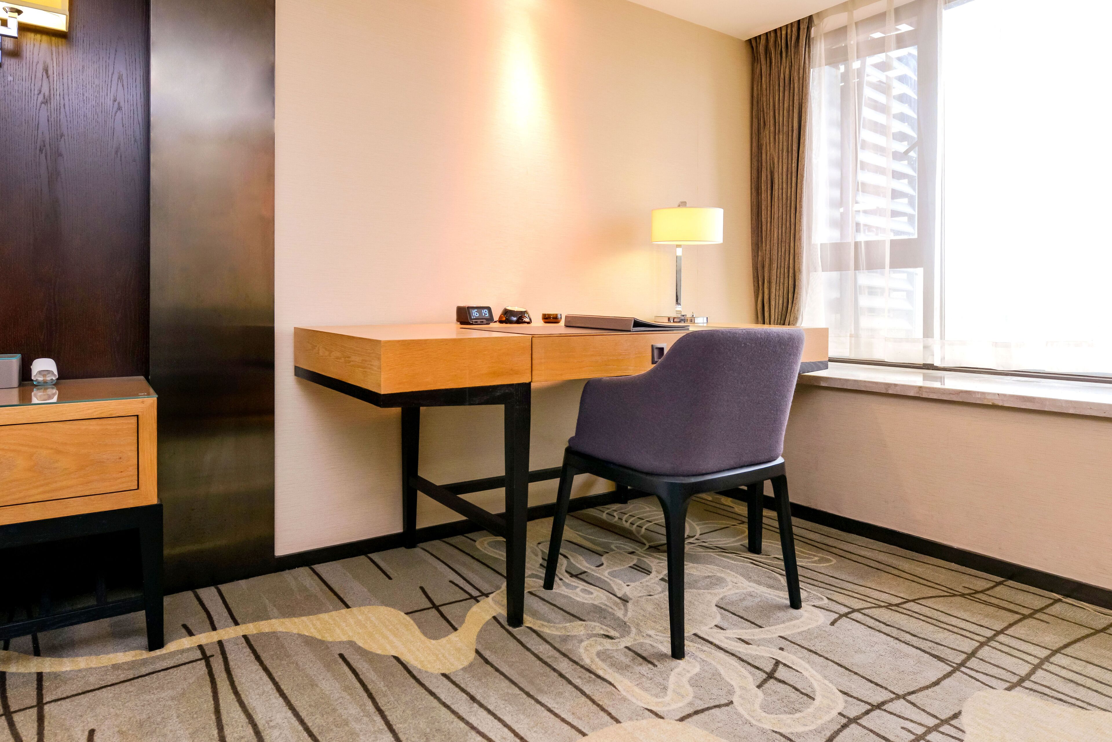 Quarto de hotel com escrivaninha de madeira, uma cadeira, abajur e janela com cortinas.