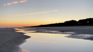 A beach with a calm water body reflecting the sky, a sandy shore, and a distant tree line. - Spacious 2-suite Condo 5-Minute Walk to the Beach. (Hilton Head Island)