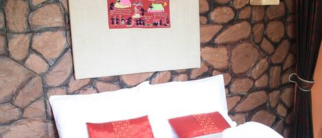A neatly made bed with white and red pillows, a remote control on a wooden side table, and a stone wall with a framed picture.