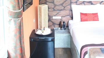 A hotel room with a stone wall, a bed with a red pillow, a small table with a kettle, and a wall-mounted air conditioner.
