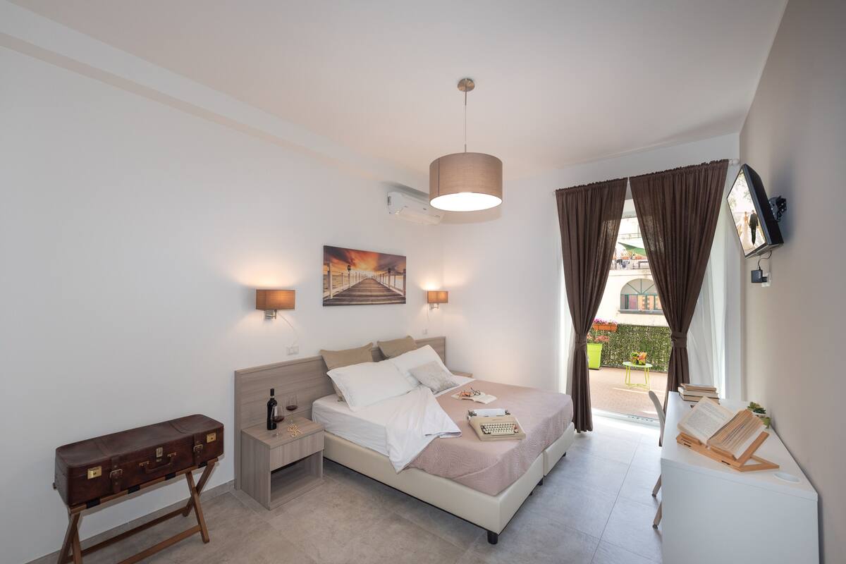 Un dormitorio con cama, mesitas de noche, escritorio, lámpara, un cuadro y vistas al exterior.