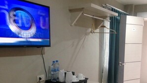 Habitación de hotel con televisor de pantalla plana montado en la pared, un refrigerador pequeño, un escritorio con bandeja de artículos de aseo y un perchero con cortina verde.