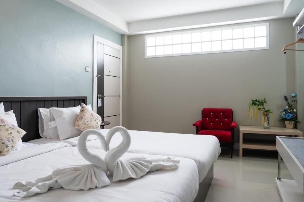 Una habitación de hotel con cama, una silla roja, una mesita de noche con un jarrón de flores y una ventana.
