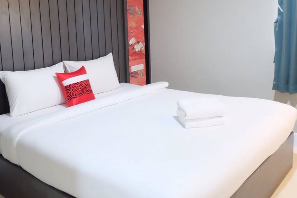 Un lit bien fait, avec une literie blanche et un oreiller rouge, une tête de lit sombre et un panneau décoratif rouge sur le mur.