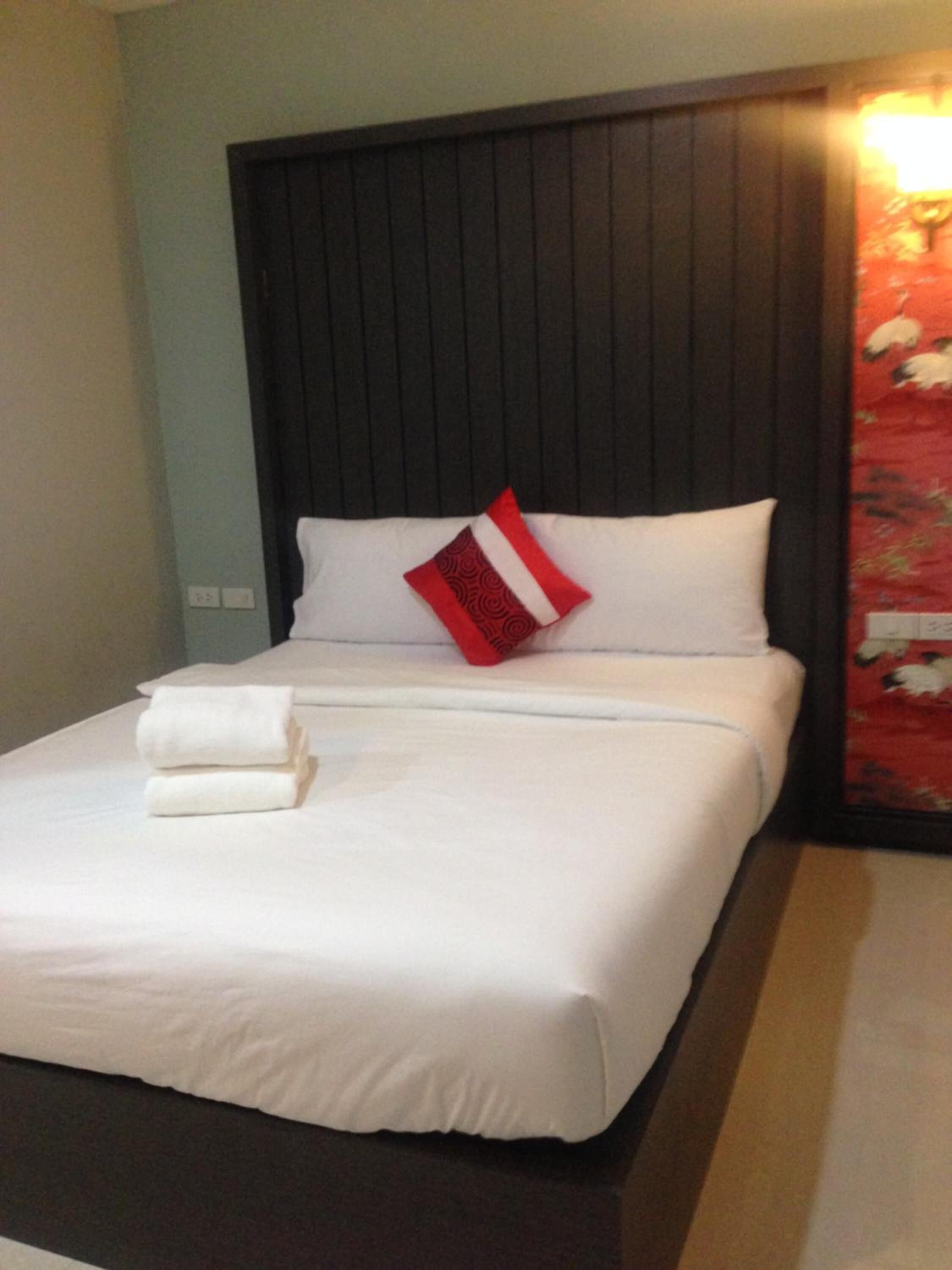A neatly made bed with white bedding and a red and white pillow, a folded white towel, and a dark wooden headboard.