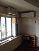 A room with a window, an air conditioner, a radiator, and a wooden shelf with items.